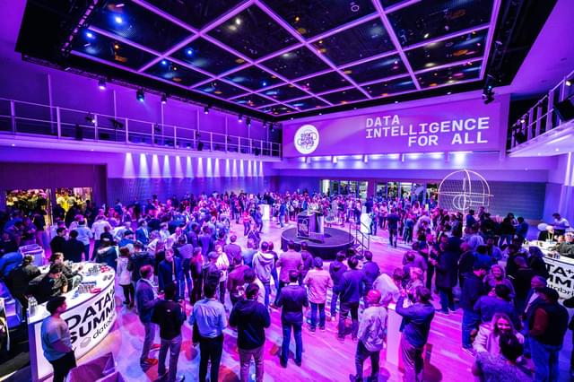 Large room with purple lighting full of people gathered around a circular stage with a person speaking.