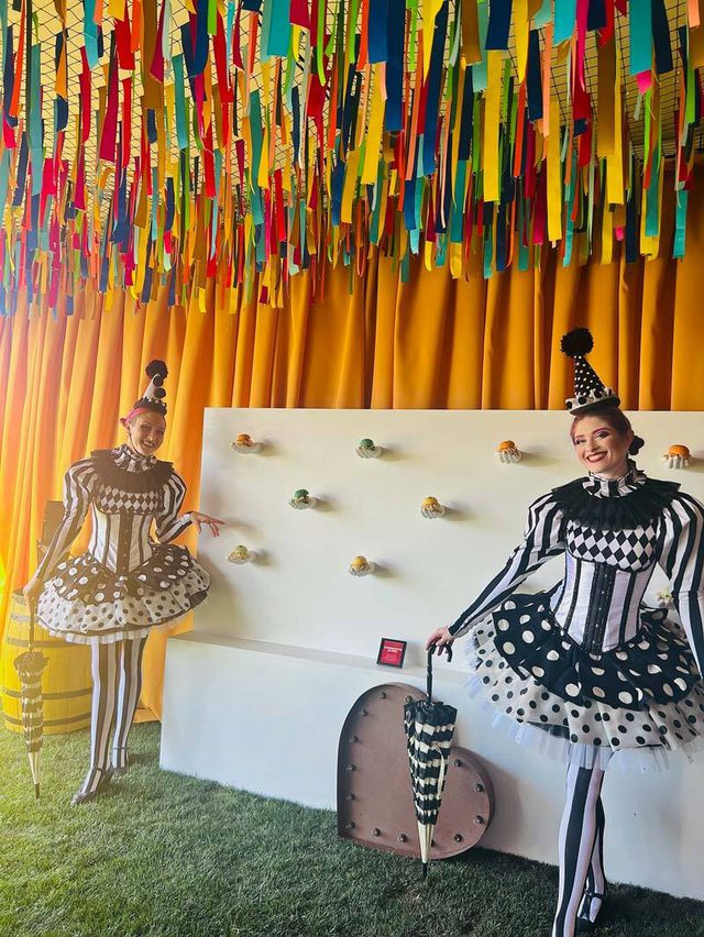 Two people in black and white circus-themed costumes in front of a display wall with sliders.