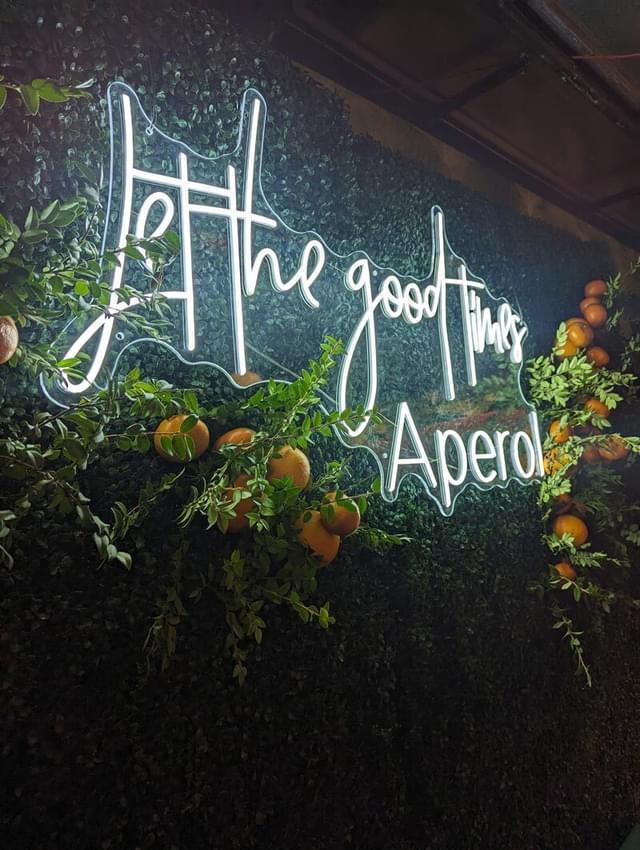 White neon sign reading "Let the good times aperol" hung on a wall with green leaves and grapefruits.