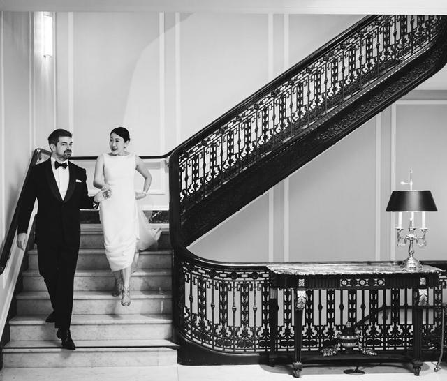 A black and white image of a couple walking down a curved staircase with a black ornate railing.