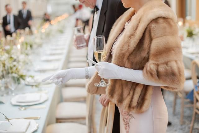 Person in a fur coat and long white gloves holding a champagne flute at a wedding reception.