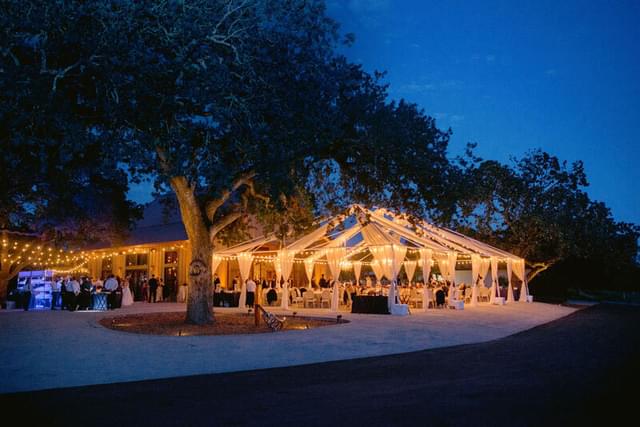 View of an outdoor wedding ceremony at dusk with white fabric draped tents and lighting.