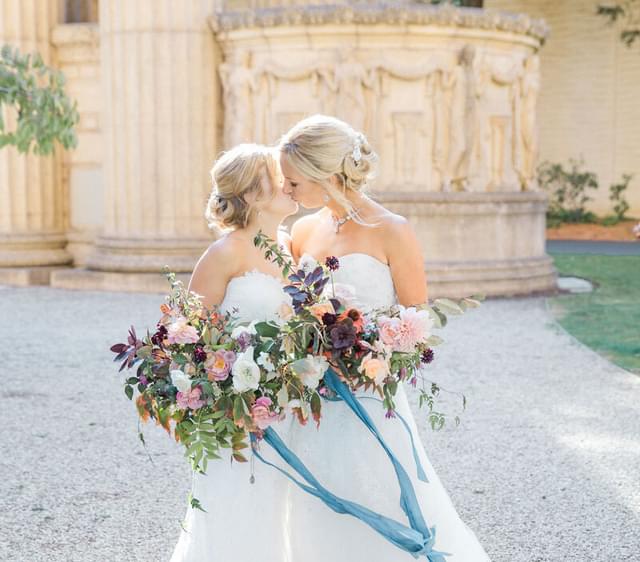 Two brides in white dresses kissing and holding colorful flower bouquets.